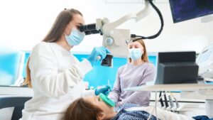 Dental professional performing a baby root canal procedure on a child under a microscope, with another staff member assisting in a pediatric dental office setting.