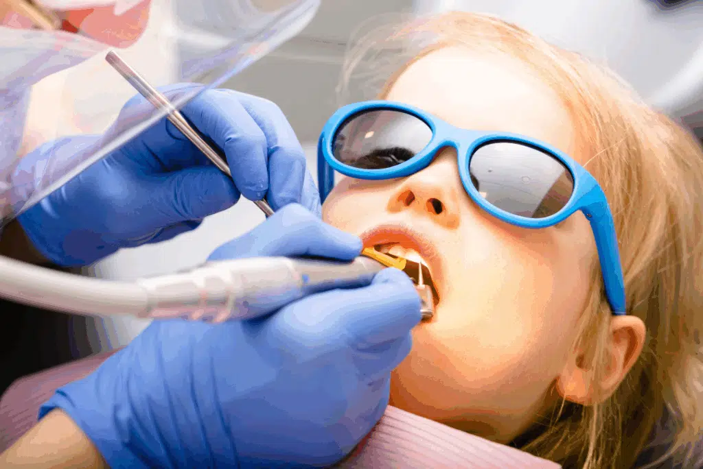 Pediatric Dentist In Great Neck, Ny: A Parent’s Guide To Expert Dental Care For Kids Child wearing sunglasses during a pediatric dental exam, with a dental professional using tools for preventive care.