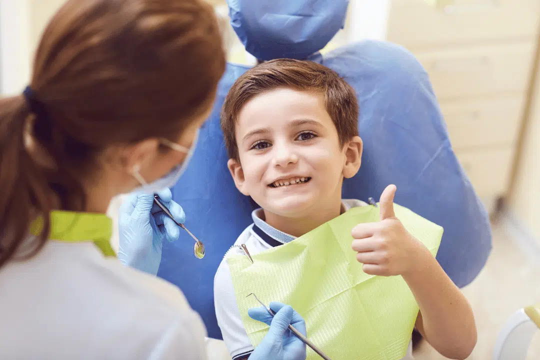 Sedation Dentistry Safety For Children: What Every Parent Should Know Child in dental chair giving thumbs up, smiling, with dentist preparing for sedation dentistry, highlighting comfort and safety in pediatric dental care.